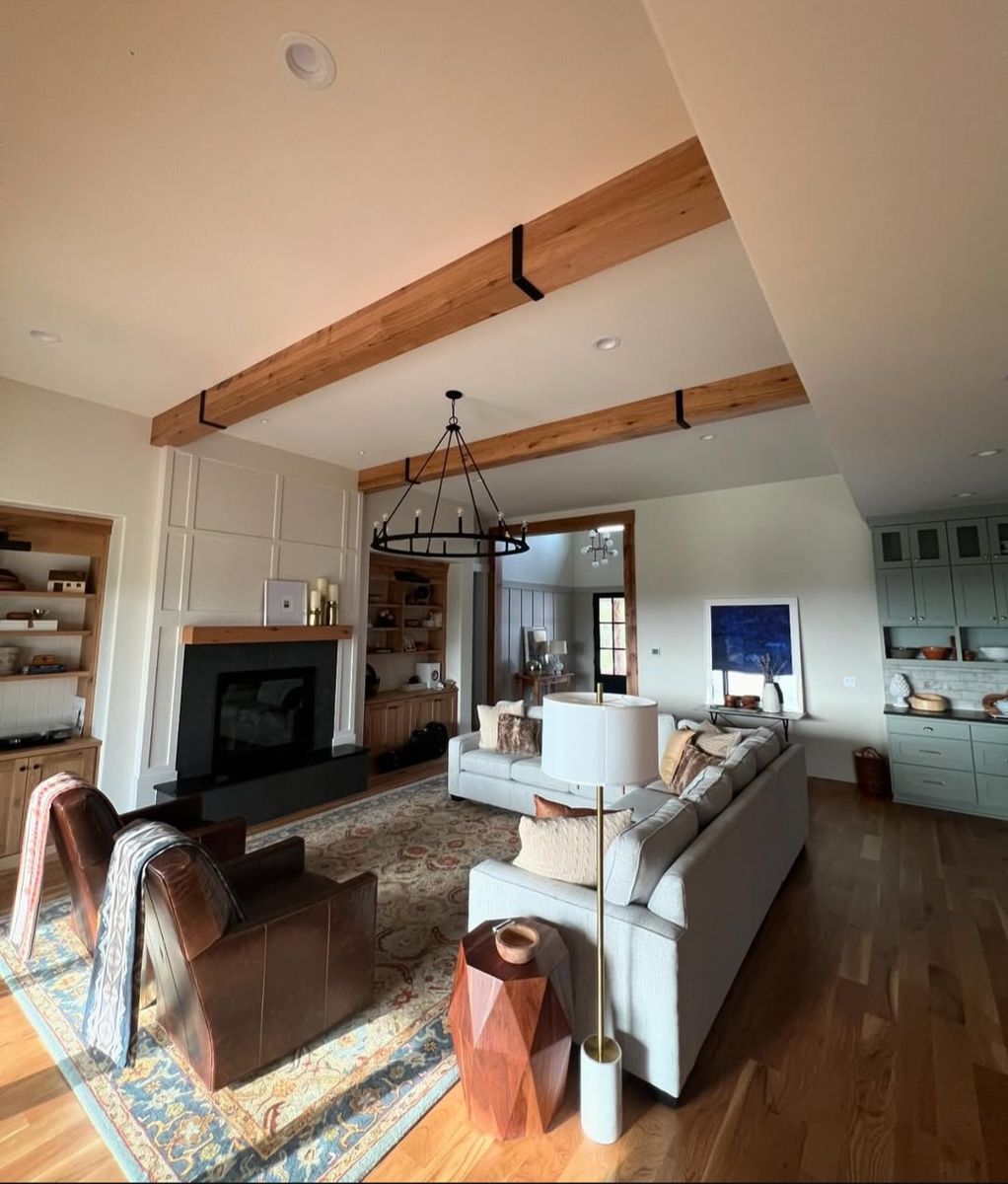 Open great room with three exposed timber beams on straps, a black slab fireplace and sage kitchen cabinetry beyond