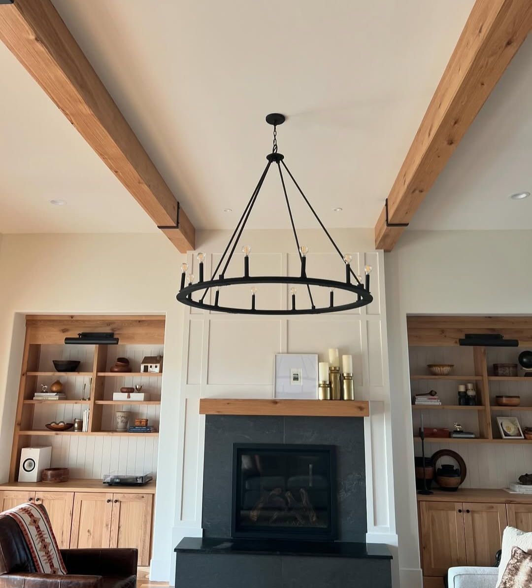 White oak built-ins flanking a panelled fireplace surround under a black wagon-wheel chandelier and exposed beams